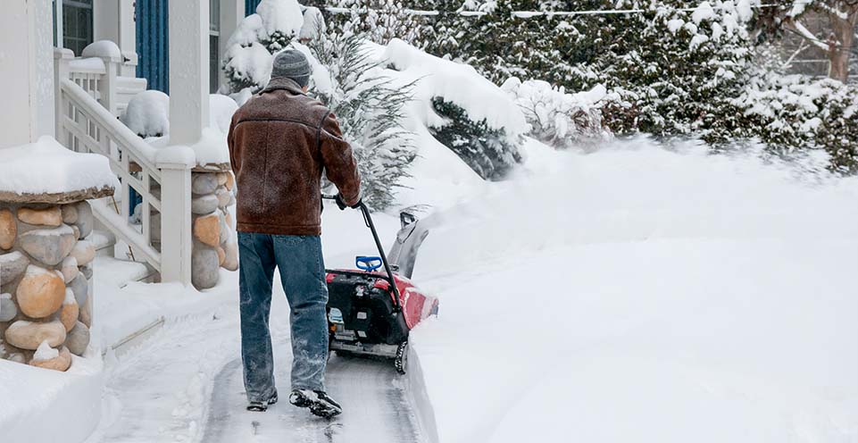Man is using a handheld snowplow to rid his home's walkway of deeply covered snow.