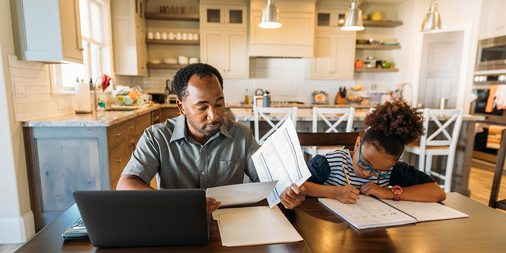 Man looking at papers and computer as child sits next to him with paper and pencil.