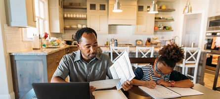 Man looking at papers and computer as child sits next to him with paper and pencil.