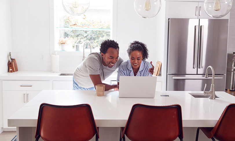 Smiling man and woman standing intently over a computer, placed on a kitchen counter.