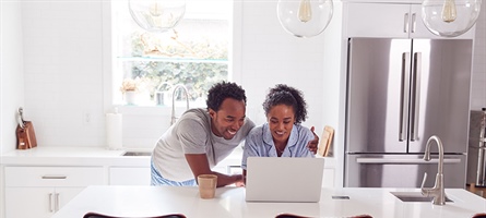Smiling man and woman standing intently over a computer, placed on a kitchen counter.