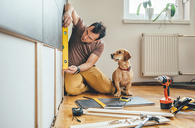 Man is seen measuring a wall for a home improvement project, while his dog sits next to him.