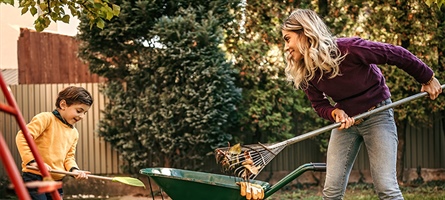 Woman and boy in a yard, holding rakes as they pile leaves into a wheelbarrow