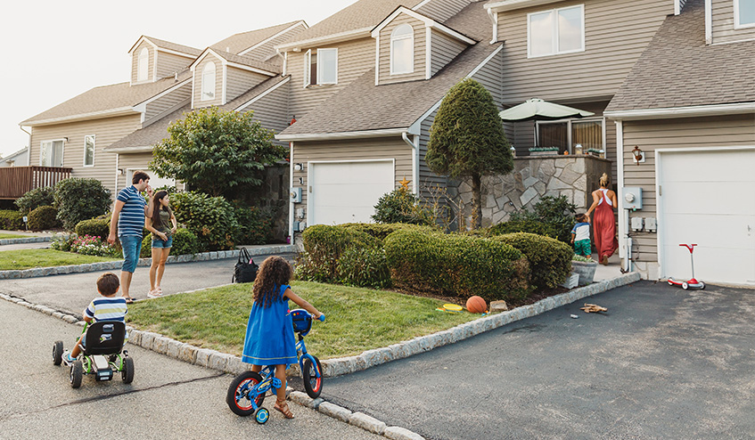 Family of two adults and two children stand outside row of houses, in front of driveway