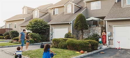 Family of two adults and two children stand outside row of houses, in front of driveway