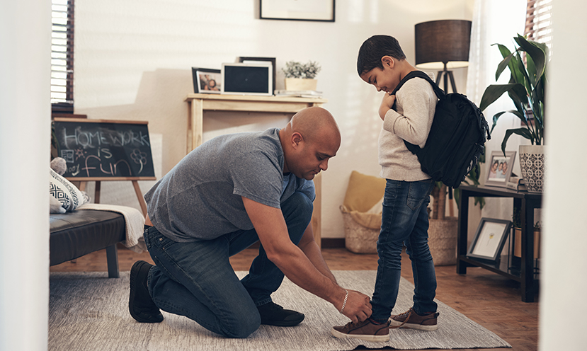 Man seen helping to tie the shoes of child with backpack, apparently headed to off to school.