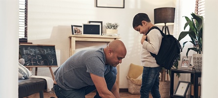 Man seen helping to tie the shoes of child with backpack, apparently headed to off to school.