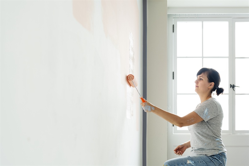 woman is kneeling while using a paint roller to paint a white wall with pink paint color