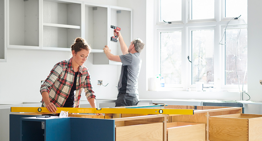 Woman measuring unfinished cabinetry and man using drill on other cabinets in the background