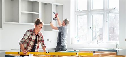 Woman measuring unfinished cabinetry and man using drill on other cabinets in the background