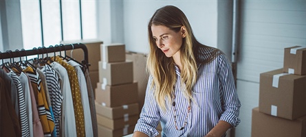 woman is surrounded by boxes and clothing as she stands to work at a computer