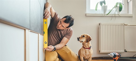 Man is seen measuring a wall for a home improvement project, while his dog sits next to him.